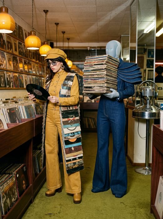 Irina Raicu in a yellow patchwork suit holding a record beside a robot mannequin carrying a stack of vinyl in a record shop
