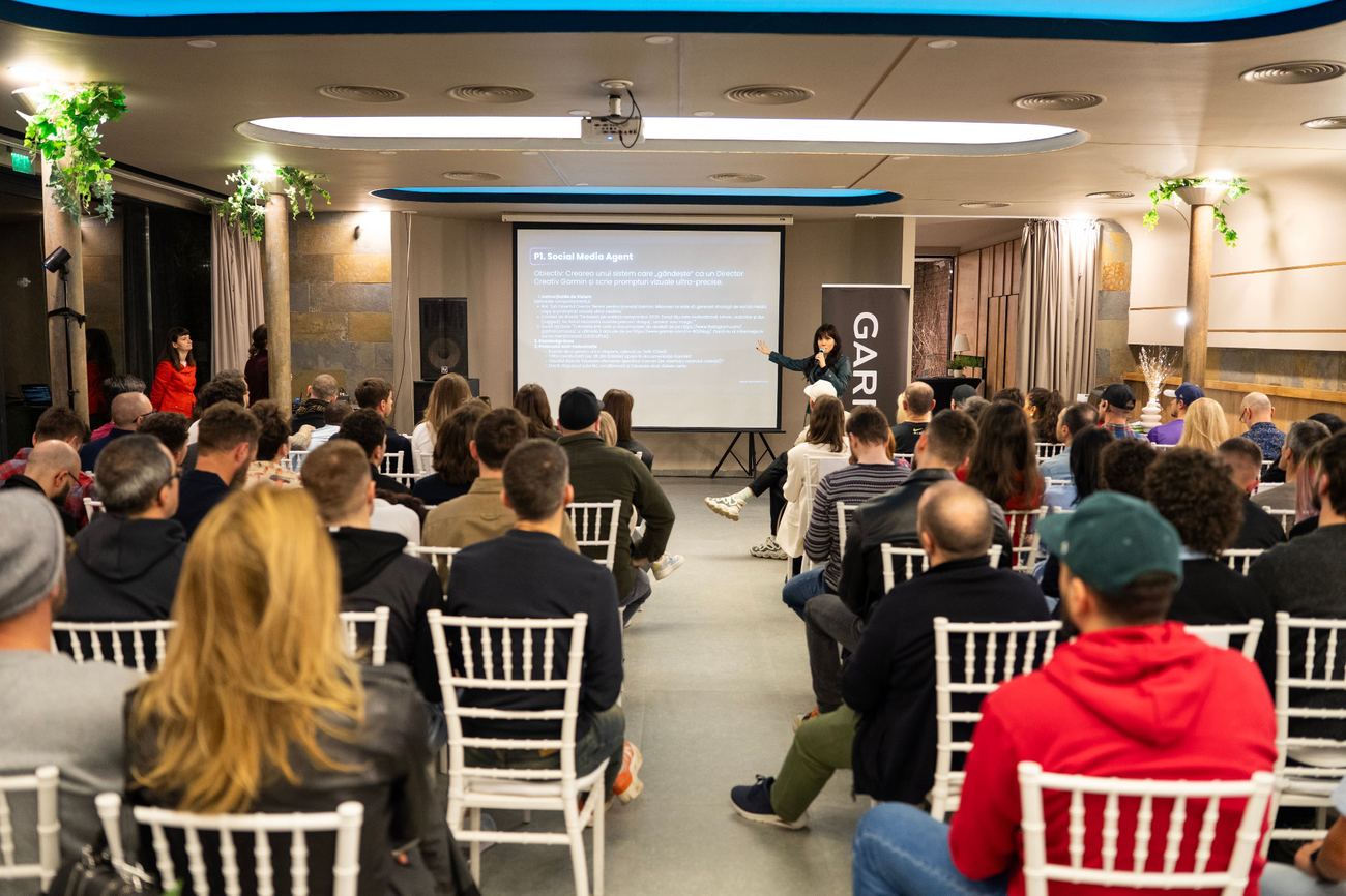 Dr. Irina Raicu speaking with a handheld microphone at the front of a room of seated attendees, beside a Garmin banner and a projected slide about a social media agent