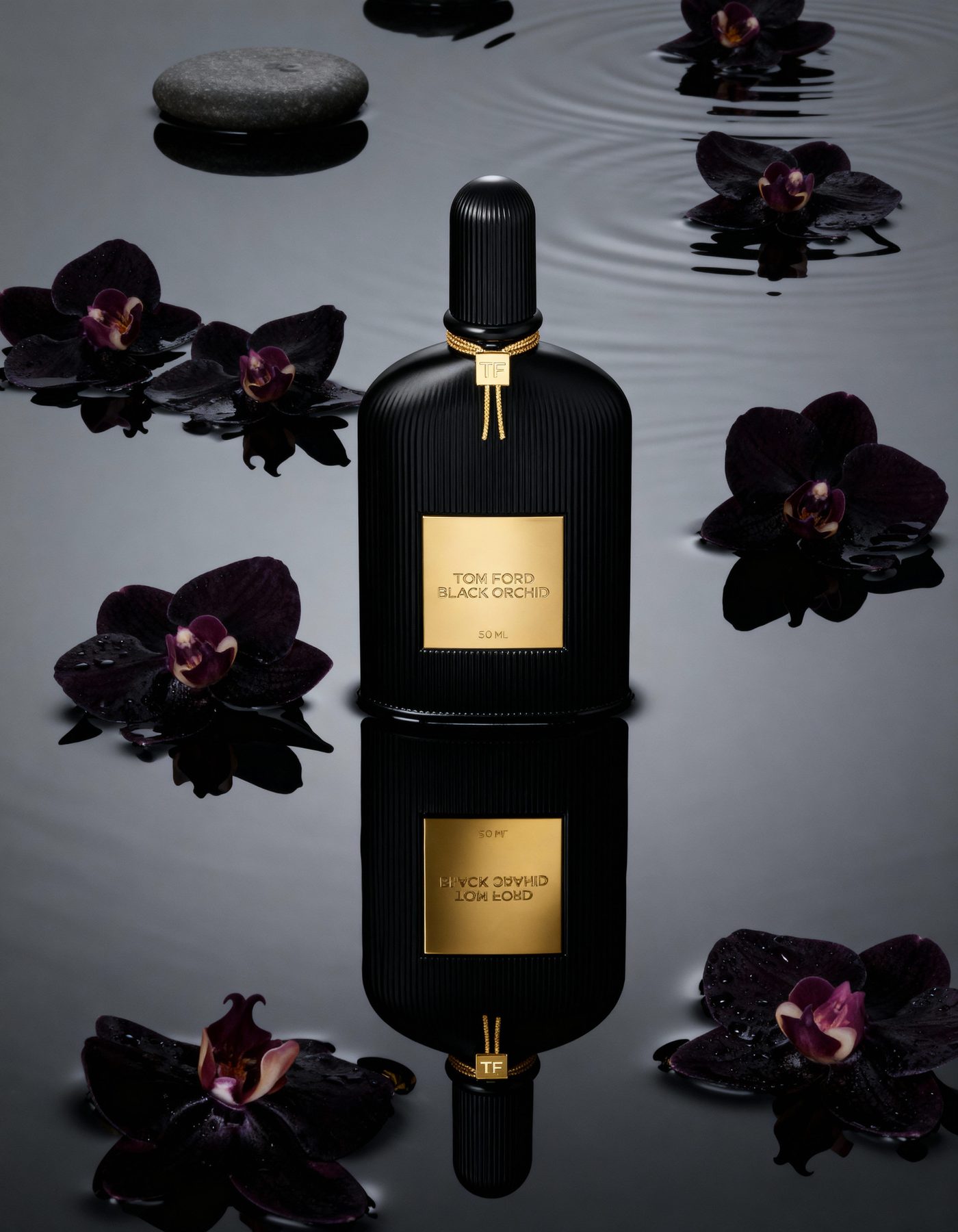 A black fragrance bottle mirrored on still water, dark orchids floating around it