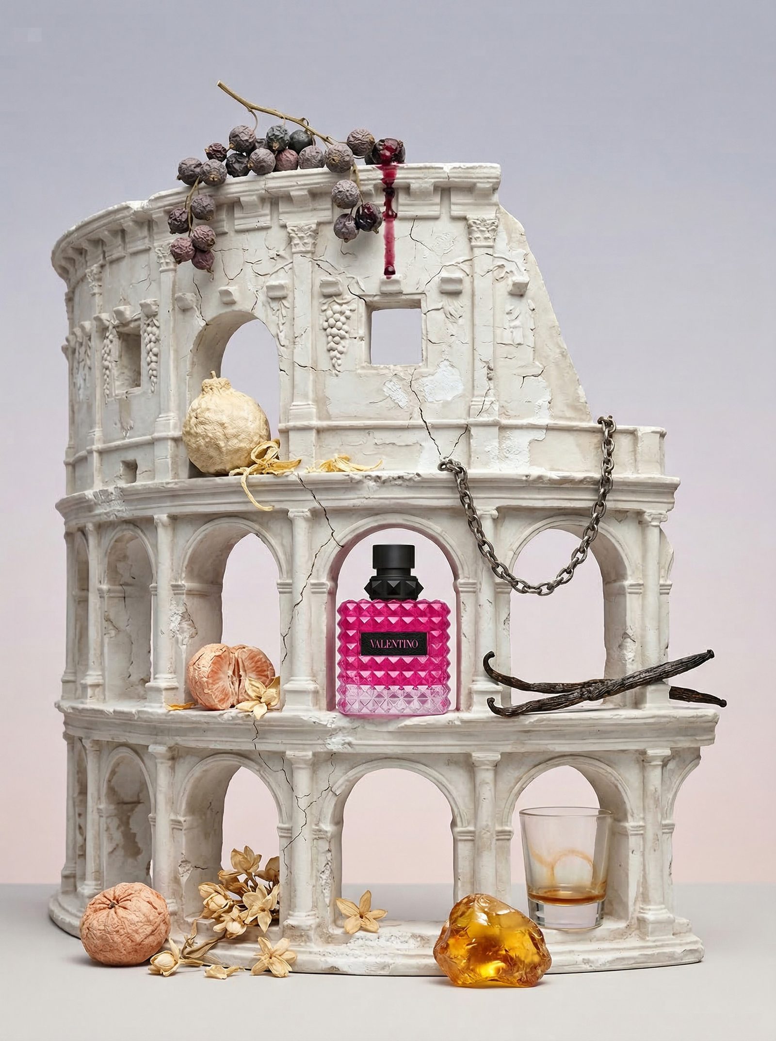 A miniature white plaster colosseum holding a pink fragrance bottle, vanilla pods and amber resin at its base