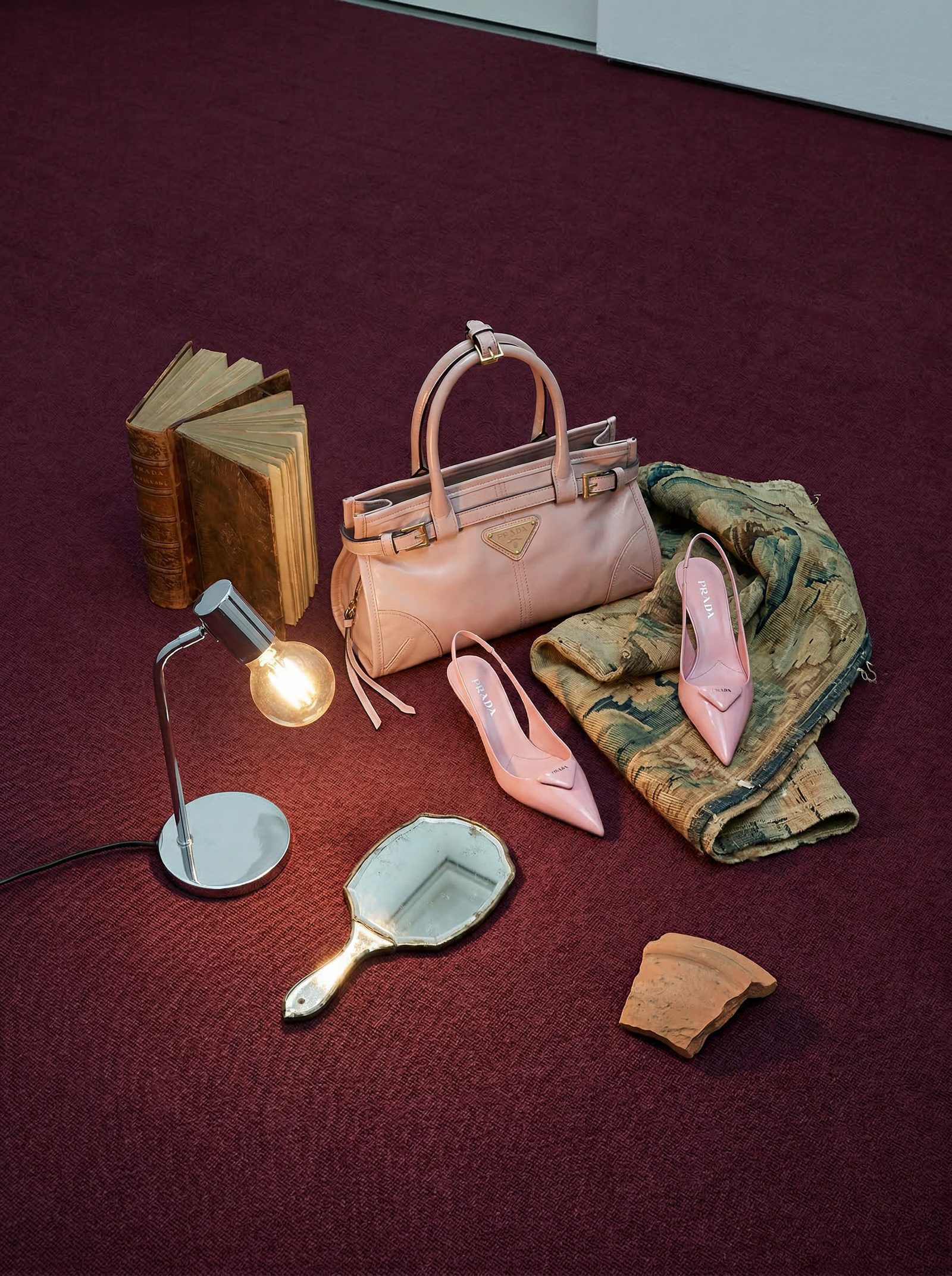 A blush pink leather handbag, heels, a hand mirror and a lit lamp arranged on deep red carpet